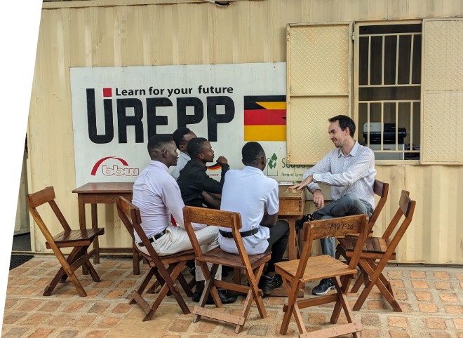 Draußen sitzen fünf junge Männer um einen Tisch und unterhalten sich. An der Wand steht ein Schild mit dem Text „Learn for your future“ und dem Projektlogo von UREPP. Holzstühle stehen um den Tisch herum. Die Szenerie strahlt eine positive Atmosphäre aus.