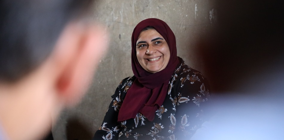 Nahaufnahme einer lächelnden Frau mit dunkelrotem Kopftuch vor unscharfem Hintergrund. Sie trägt ein dunkelbraunes, gemustertes Oberteil. Der Blickwinkel ist leicht von der Seite. Die Stimmung ist freundlich und positiv.