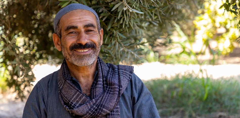 Lächelnder Mann mittleren Alters mit grauen Haaren und einem grauen Schal vor einem Olivenbaum. Er trägt eine graue Mütze. Das Bild strahlt Wärme und Ruhe aus.