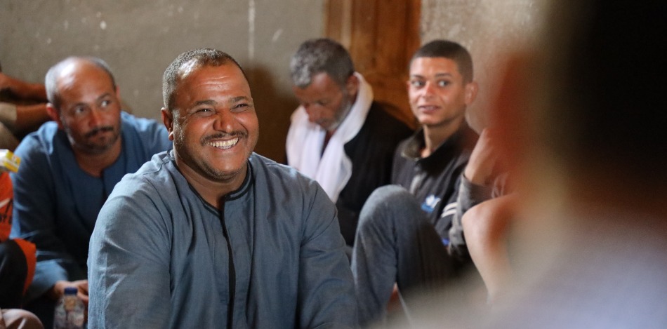 Ein lächelnder Mann im Vordergrund, umgeben von anderen Männern in einem Raum mit schlichter Einrichtung. Das Bild vermittelt ein Gefühl von Gemeinschaft und positiver Stimmung. Die Männer sitzen beisammen und scheinen sich zu unterhalten. Die Stimmung ist entspannt und freundlich.