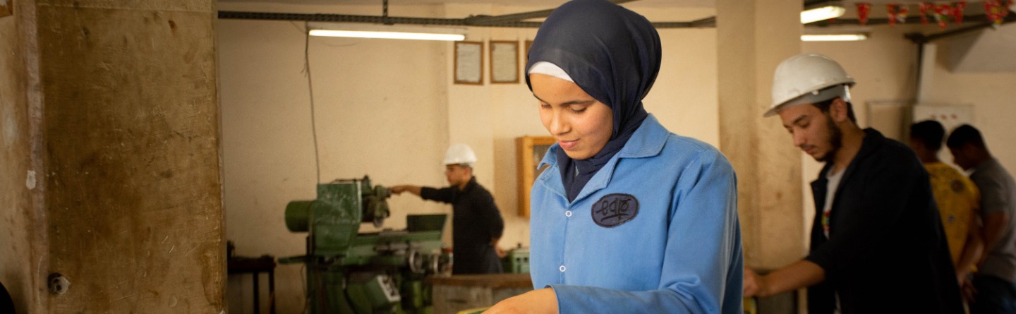 Das Bild zeigt eine junge Frau in einer Werkstatt am Schraubstock bei der Bearbeitung eines Metallteiles. 