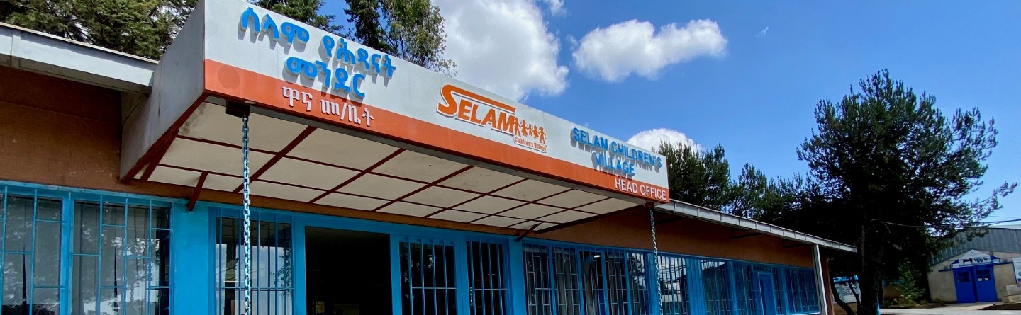 Sonniger Tag. Ein niedriges, ockerfarbenes Gebäude mit hellblauen Türen und Fenstern. Ein Schild weist auf das "Selam Children's Village Head Office" – den Eingangsbereich des Colleges hin. Pflanzen schmücken den Eingangsbereich.