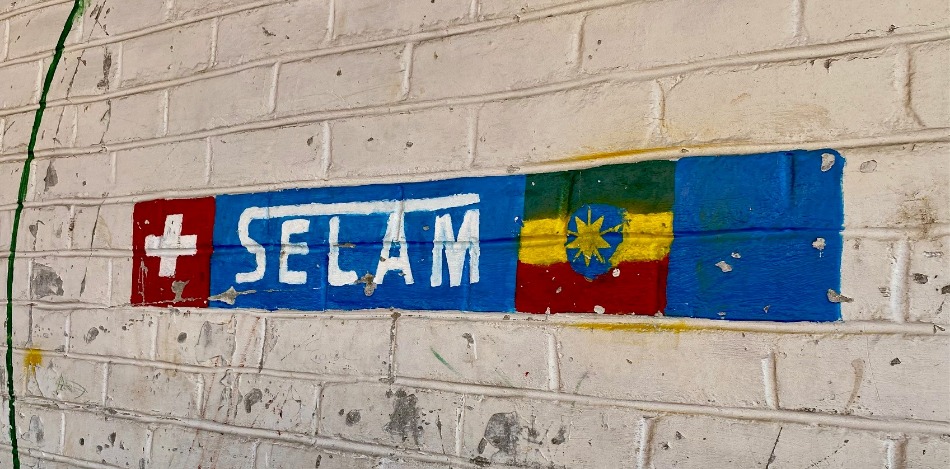 Auf einer weißen Backsteinmauer ist ein Wandgemälde zu sehen. Es zeigt die Schweizer Flagge, das Wort „SELAM“ (Frieden auf Amharisch) und die äthiopische Flagge. Die Farben sind blau, rot, gelb, grün und weiß. Das Bild vermittelt einen Eindruck von Internationalität und Frieden.