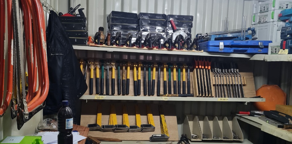 Eine gut sortierte Werkbank in einem Container. Es gibt zahlreiche Werkzeuge, wie Sägen, Hämmer und Maßstäbe, ordentlich in Regalen verstaut. Auf dem Tisch liegen weitere Werkzeuge. Der Raum ist beleuchtet durch eine Neonröhre.