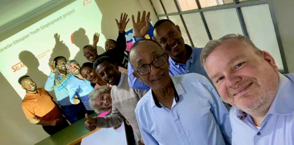 Gruppenfoto der Teilnehmer*innen des Planungsworkshops 2024. Mehrere Menschen lächeln in die Kamera, ein Mann macht ein Selfie. Die Szenerie ist ein Konferenzraum. Ein Projektor zeigt "Bayarian Ethiopian Youth Employment Project".