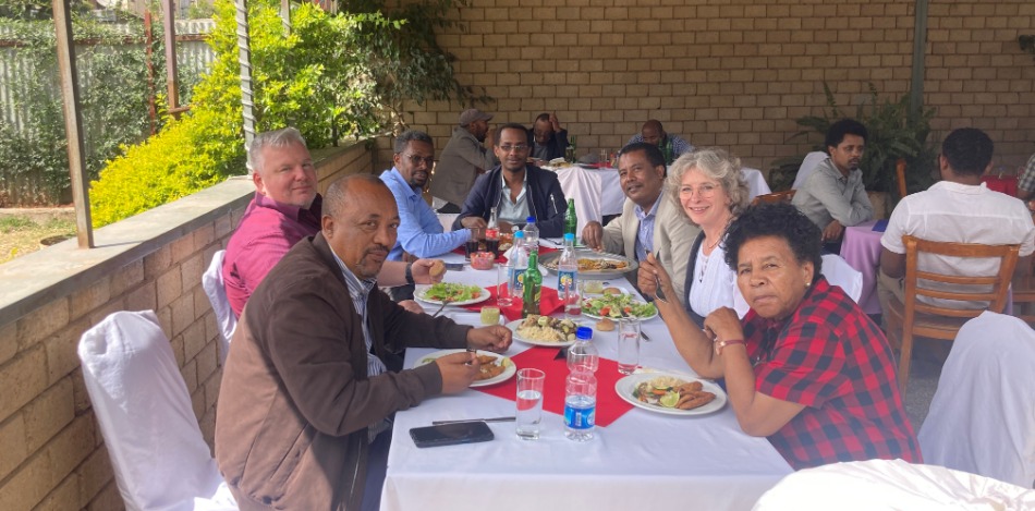 Hier genießen mehrere Personen ein gemeinsames Essen im Restaurant des Selam Colleges. Angeordnet an Tisch und Stühlen, sind sie in angeregter Stimmung. Sonnenlicht fällt auf die Szene.