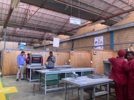 Ein heller Werkraum mit mehreren Arbeitstischen, Werkzeugen und Geräten. Studenten in Arbeitskleidung arbeiten an Projekten. Ein Schild zeigt „Demonstrationsbereich“ an. Der Raum ist gut beleuchtet und geräumig.