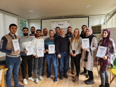 Zwölf Personen, die meisten Männer, halten Zertifikate. Eine Frau steht im Zentrum. Hintergrund: Seminarraum. Thema: Weiterbildung/Abschluss.