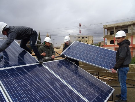 Vier Personen in weißen Helmen montieren unter bewölktem Himmel Solarmodule auf einem Dach. Der Fokus liegt auf nachhaltiger Energiegewinnung.