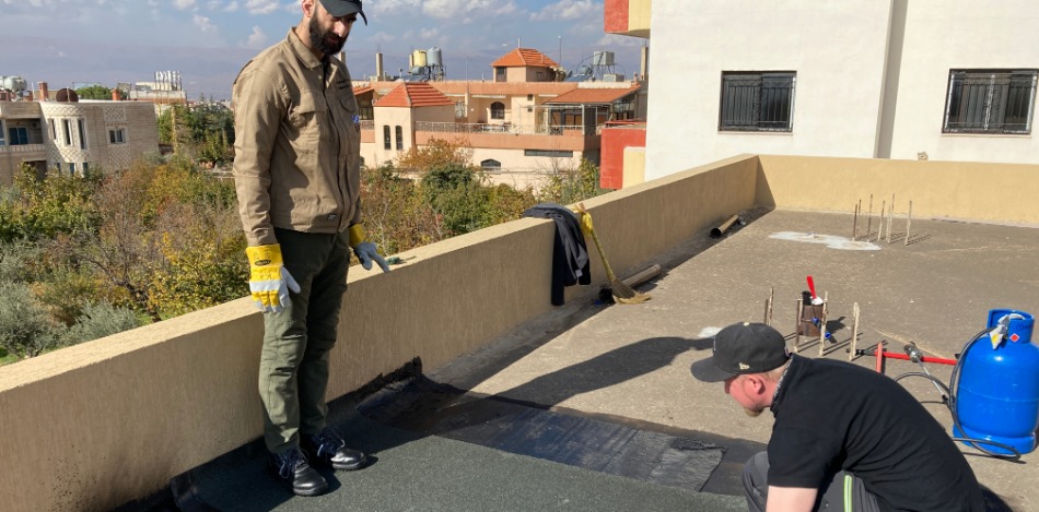 Anbringung von zwei Lagen Bitumenbahnen auf einem begehbaren Flachdach. Arbeiten erfolgen im Rahmen eines Trainingszwecks.