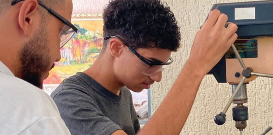 Das Bild zeigt zwei Jugendliche an der Bohrmaschine, wo sie ein Loch in die Erkennungsmarke bohren. 