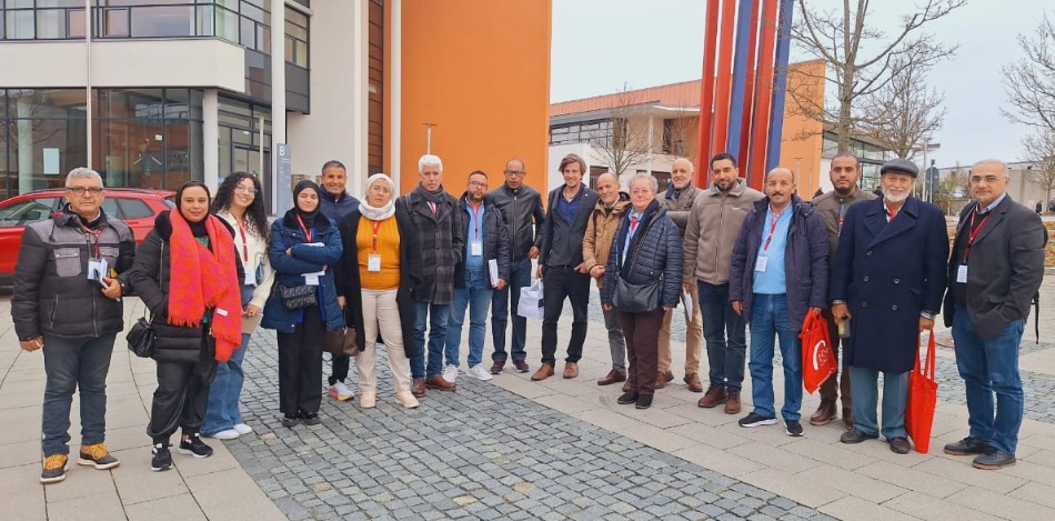 Eine Gruppe von etwa 15 Personen, hauptsächlich Männer, posiert vor einem Gebäude. Sie tragen unterschiedliche Kleidung. Einige halten rote Taschen. Im Hintergrund ist ein Gebäude und ein modernes Kunstwerk zu sehen.