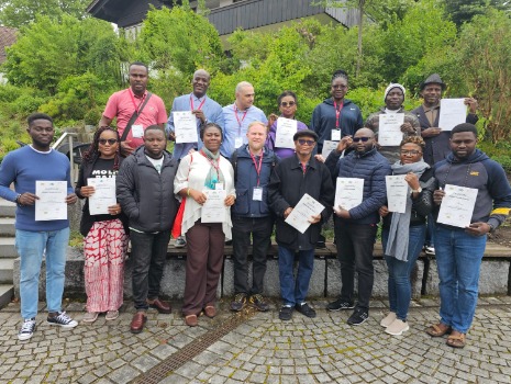 Gruppe von 16 Personen, überwiegend afrikanisch, hält Zertifikate in den Händen. Sie stehen auf einem gepflasterten Platz vor einem Gebäude und Grün. Freude und Erfolg sind erkennbar.