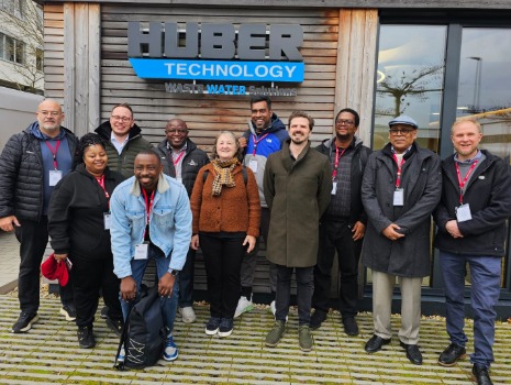 Vor dem Huber Technology Gebäude stehen 11 Personen lächelnd in zwei Reihen. Sie tragen Jacken und Konferenz-Ausweise. Die Gruppe ist multiethnisch. Das Gebäude ist aus Holz.