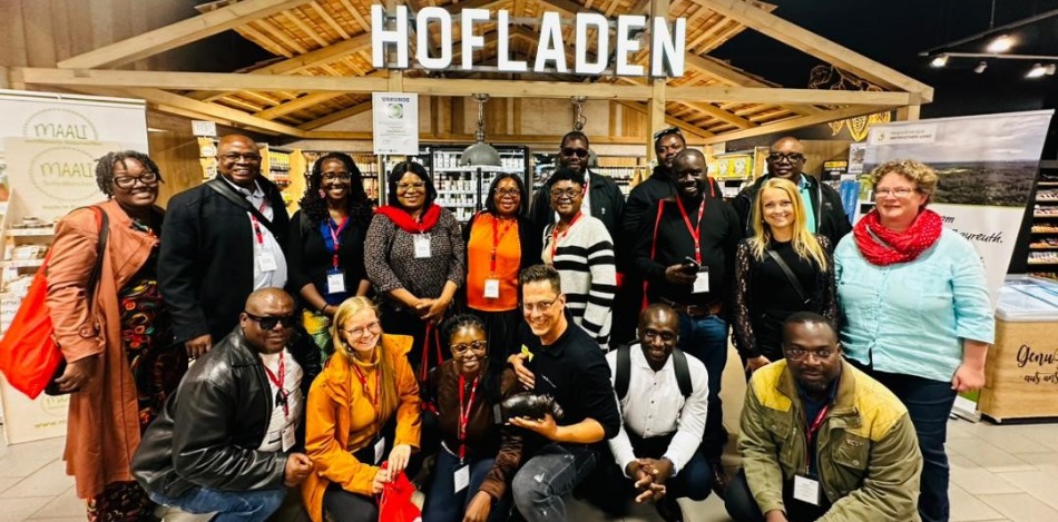 Große Gruppe lächelnder Menschen vor einem Hofladen-Stand. Unterschiedliche Altersgruppen und Ethnien sind vertreten. Freundliche, einladende Atmosphäre.