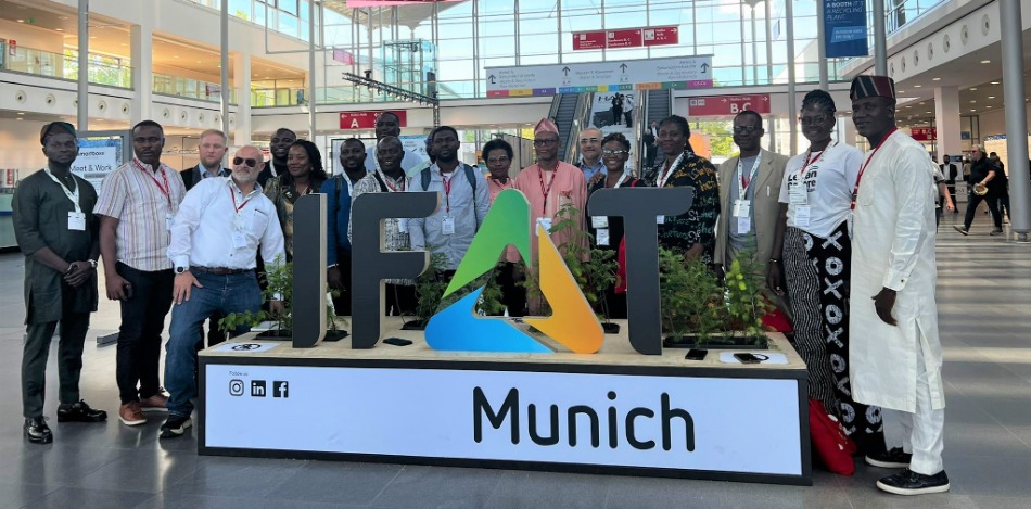Gruppenfoto vor dem IFAT-Logo in München. Eine diverse Gruppe von Menschen posiert vor einem großen Buchstaben-Logo mit Pflanzen und einem Bildschirm im Hintergrund.