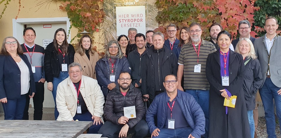 Eine Gruppe von etwa 20 Personen posiert vor einem Gebäude. Ein Schild wirbt für den Ersatz von Styropor. Herbstlaub umrahmt das Bild. Die Personen tragen unterschiedlich farbige Kleidung und Ausweise. Sie lächeln in die Kamera.