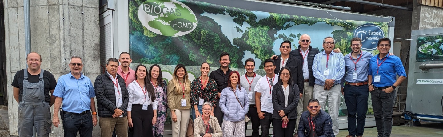 Eine Gruppe von ca. 20 Personen steht vor einem Biogas-Container. Sie lächeln in die Kamera. Der Container zeigt ein Weltkartenmotiv. Das Foto wirkt freundlich und professionell.