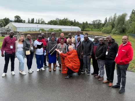 Eine Gruppe von etwa 20 Personen posiert für ein Foto im Freien, mit einem Mann in einem gemusterten Umhang, der kniet und die Arme ausstreckt. Im Hintergrund sind Gebäude und Bäume zu sehen.