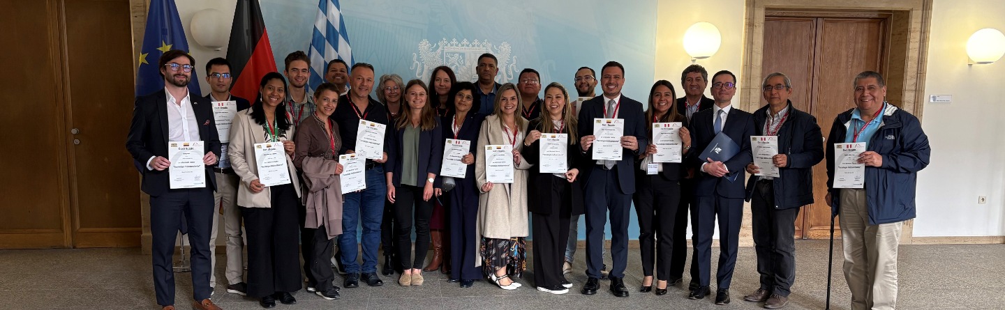 Eine Gruppe von Personen posiert mit Urkunden vor Flaggen und Wappen. Die meisten lächeln.