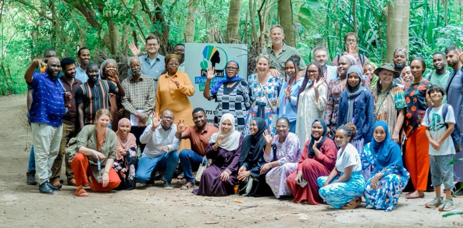 Eine Gruppe von etwa 40 Personen posiert für ein Foto vor einem grünen Blätterdach, viele winken. Ein Schild "Practical Permaculture Institute" ist sichtbar.