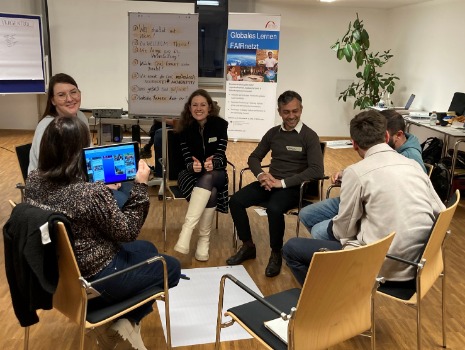 Bild zeigt eine Gruppe von Menschen, die in einem Kreis auf Stühlen sitzen und sich unterhalten. Ein Flipchart und ein Tablet sind sichtbar. Die Atmosphäre wirkt freundlich und kollegial. Ein Workshop oder Meeting.