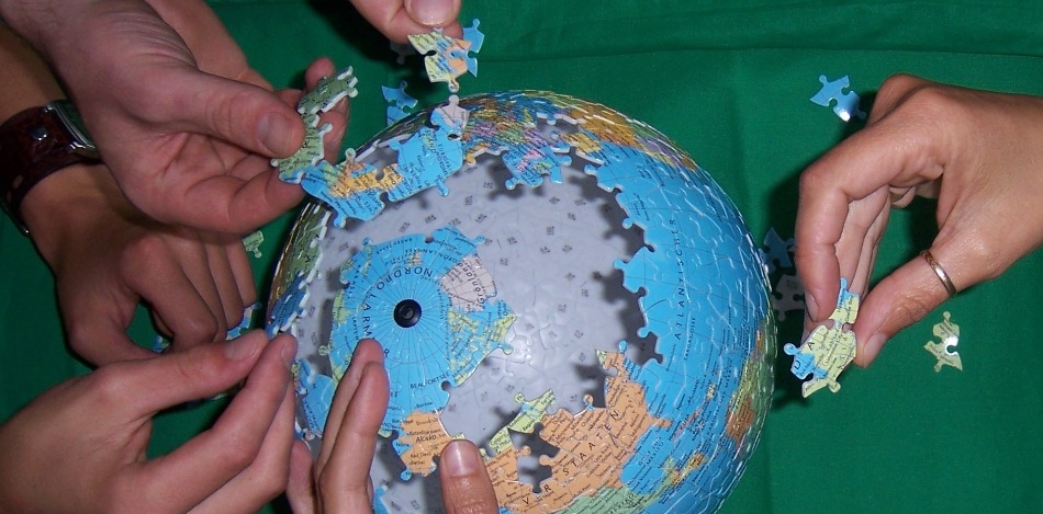 Mehrere Hände setzen ein unvollständiges Globus-Puzzle zusammen. Die Teile liegen auf grünem Untergrund. Das Bild symbolisiert globale Zusammenarbeit.