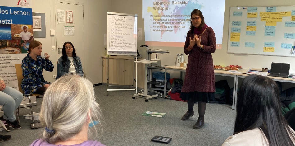 Eine Frau hält einen Vortrag über "Lebendige Statistik: Weltspiel" vor einer Gruppe von Zuhörern. Folien zeigen Weltbevölkerung, BIP-Verteilung und Flüchtlingszahlen. Ein entspanntes, informatives Setting.