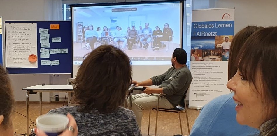 Ein Mann sitzt und präsentiert eine Videokonferenz mit einer Gruppe von Menschen auf einem Bildschirm. Das Thema scheint globales Lernen zu sein. Im Raum sitzen weitere Personen und verfolgen die Präsentation. Ein Flipchart mit Notizen steht im Hintergrund.