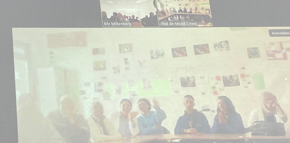 Bild zeigt einen Screenshot einer Videokonferenz. Eine Gruppe junger Menschen sitzt an einem Tisch vor einer Wand mit Bildern. Im Hintergrund weitere Teilnehmer. Der Bildschirm wirkt unscharf.