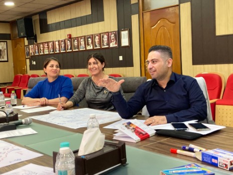 Drei Personen sitzen an einem Tisch und diskutieren bei einem Feinplanungsworkshop. Auf dem Tisch liegen Unterlagen, Stifte und Wasserflaschen. Im Hintergrund hängen Porträts an der Wand. Ein produktives und kollaboratives Arbeitsumfeld.