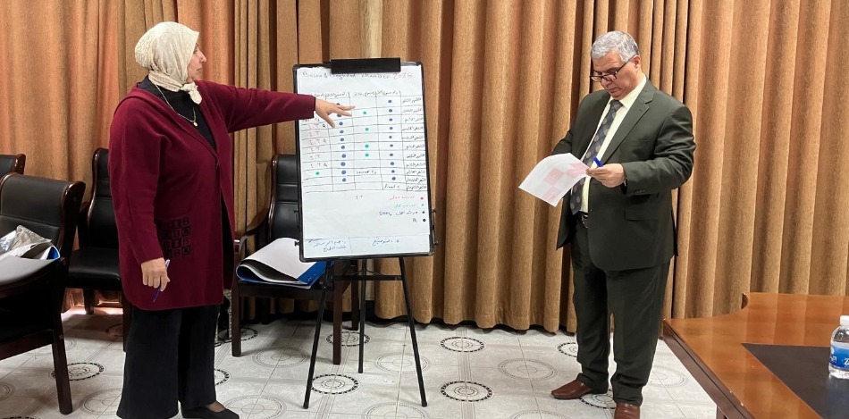 Zwei Personen, eine Frau in einem roten Cardigan und ein Mann im Anzug, besprechen während des Planungsworkshops 2025 in Basra eine Tabelle an einem Flipchart. Der Raum ist mit braunen Vorhängen ausgestattet.