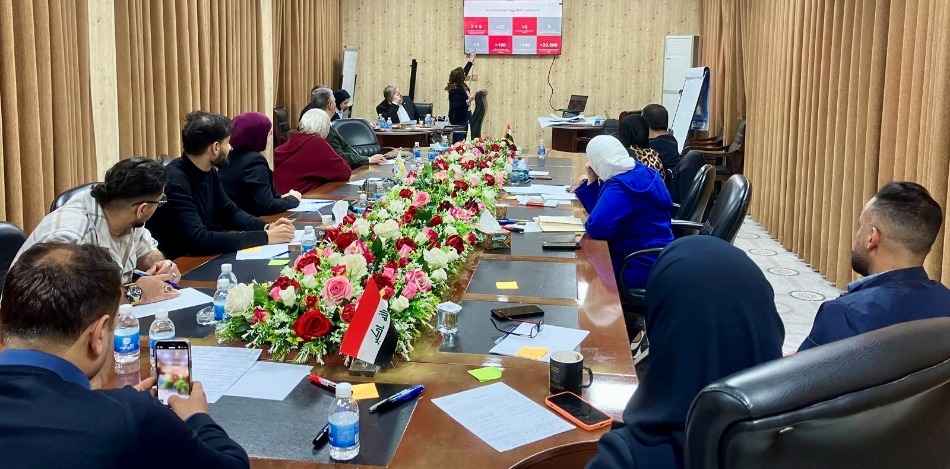 Hier tagt ein großer Kreis von Personen aus den Kammern von Basra und Bagdad um einen langen Tisch. Ein Blumenarrangement und eine irakische Flagge zieren den Tisch. Eine Frau präsentiert etwas an einer Leinwand.
