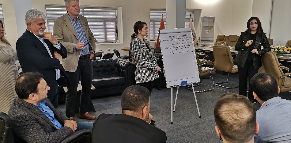 Gruppenfoto eines Workshops in Erbil zu Fachgruppen. Eine Frau präsentiert an einem Whiteboard. Teilnehmer*innen sitzen und stehen im Raum. Diskussion und Austausch im Fokus.