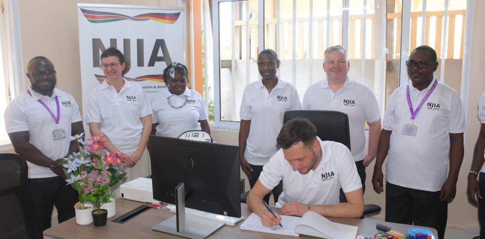 Mehrere Mitglieder des NJIA-Projektteams stehen gemeinsam in einem Büro am Kenya Coast National Polytechnic. Eine Person sitzt am Schreibtisch und unterschreibt ein Dokument. Im Hintergrund ist ein NJIA-Roll-up zu sehen. Das Bild zeigt die Einrichtung und Nutzung des lokalen Projektbüros sowie die Zusammenarbeit zwischen bbw gGmbH, KCNP und weiteren Projektpartnern.
