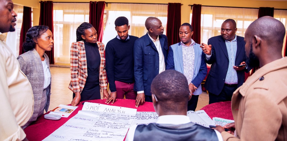Eine Gruppe von Personen steht um einen Tisch und bespricht eine SWOT-Analyse, die auf großen Plakaten notiert ist.