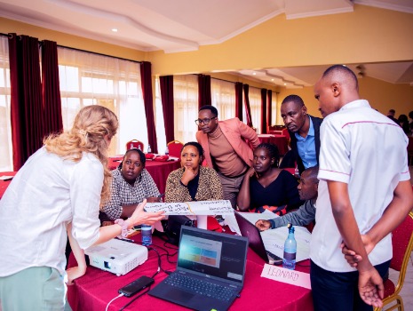 Das Bild zeigt acht Personen, die sich in einem Workshop konzentriert um einen Tisch versammelt haben und gemeinsam an Plakaten, einem Laptop und einem Projektor arbeiten.