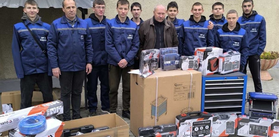 Eine Gruppe junger Männer in blauen Arbeitsanzügen steht vor einer Berufsschule, umgeben von neuen Werkzeugkoffern, Kartons mit technischer Ausrüstung und einem blauen Werkzeugschrank.