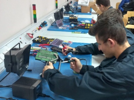 Jugendliche in dunkler Arbeitskleidung arbeiten in einem modern ausgestatteten Elektroniklabor mit Lötstationen und Leiterplatten an blauen Arbeitsmatten.