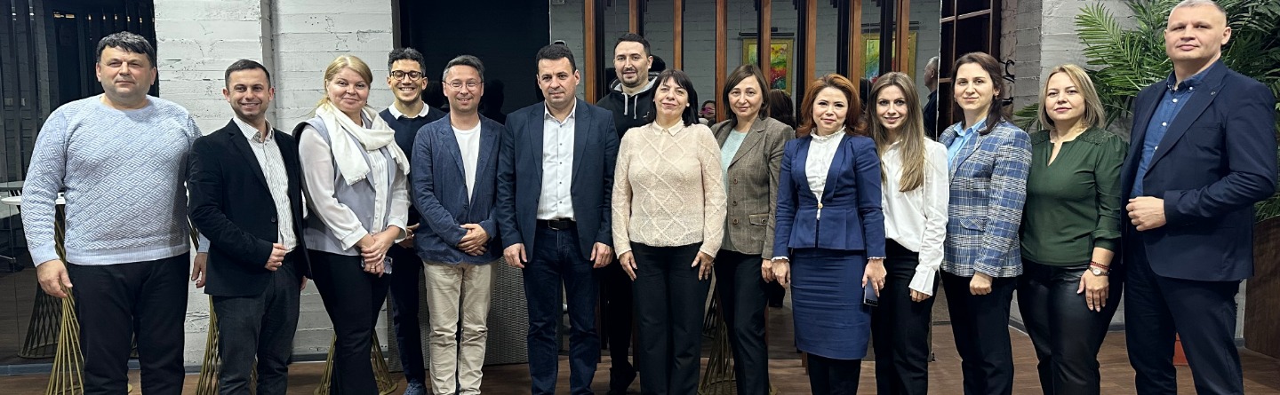 Mehrere Teilnehmer sitzen an einem langen Tisch mit Notizbüchern und Wasserflaschen, während eine Präsentation auf einer Leinwand gezeigt wird.
