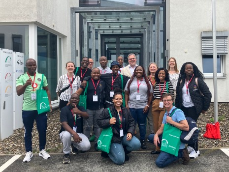 Eine Gruppe von Menschen steht vor einem Eingang in ein Gebäude in drei Reihen. Die Personen haben grüne Taschen und lachen. 