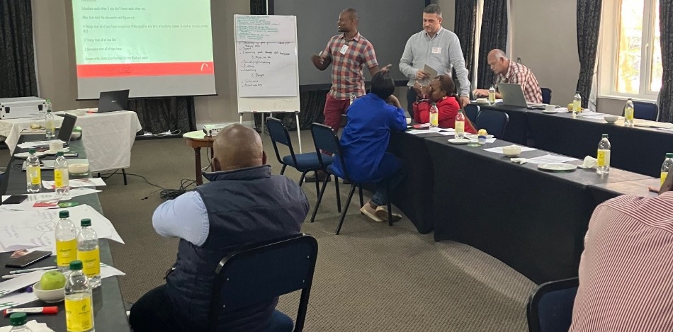 Einige Menschen sitzen und stehen in einem Seminarraum. Eine Person präsentiert am Flipchart. 