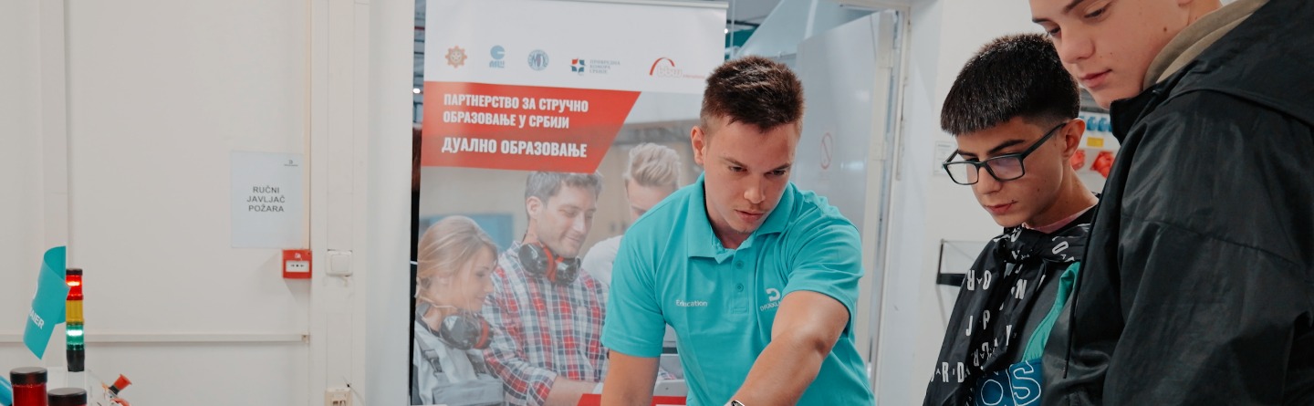 Das Bild zeigt einen Auszubildenden, der zwei Grundschülern die Technik an seinem Arbeitsplatz erklärt und ihnen praktische Anwendungen näherbringt.