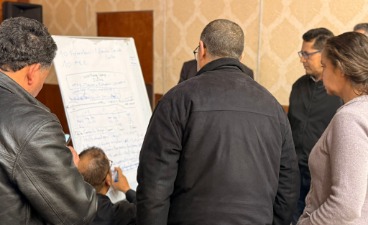Mehrere Personen stehen im Halbkreis um ein Flipchart und arbeiten gemeinsam an Inhalten. Eine Person schreibt auf das Flipchart, auf dem handschriftliche Notizen und Skizzen zu sehen sind.