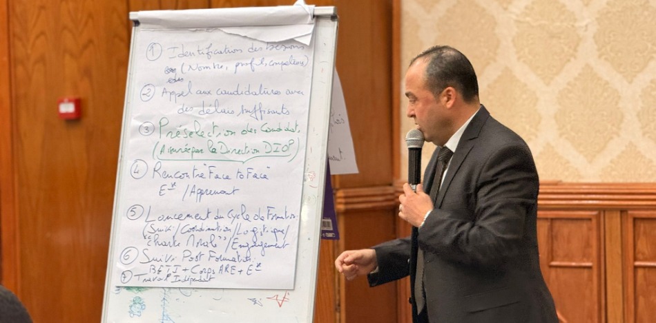 Mann im Anzug spricht in Mikrofon vor Flipchart mit Notizen.