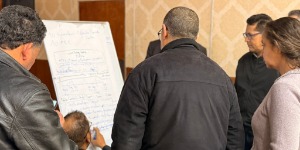 Mehrere Personen stehen im Halbkreis um ein Flipchart und arbeiten gemeinsam an Inhalten. Eine Person schreibt auf das Flipchart, auf dem handschriftliche Notizen und Skizzen zu sehen sind.