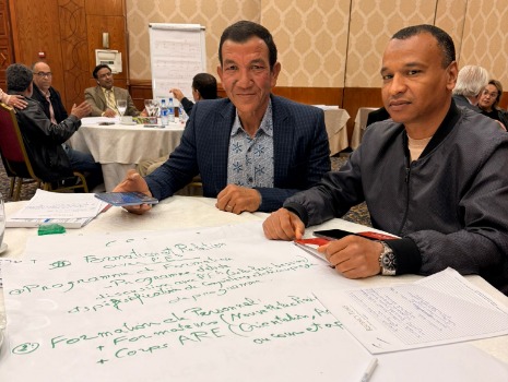 Zwei Personen sitzen an einem Tisch in einem Seminarraum und arbeiten gemeinsam. Auf dem Tisch liegen ein großes Papier mit handschriftlichen Notizen, Stifte und ein Smartphone. Im Hintergrund sitzen weitere Teilnehmende an runden Tischen und diskutieren.