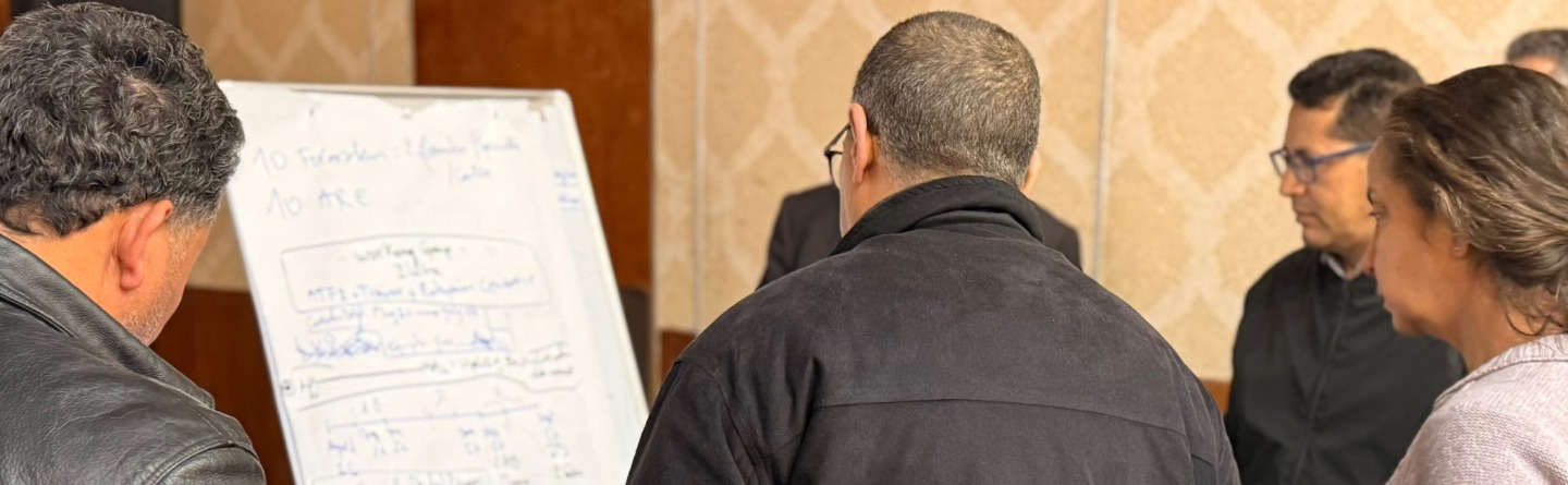 Gemeinsames Brainstormen der Projektaktivitäten Mehrere Personen stehen im Halbkreis um ein Flipchart und arbeiten gemeinsam an Inhalten. Eine Person schreibt auf das Flipchart, auf dem handschriftliche Notizen und Skizzen zu sehen sind.