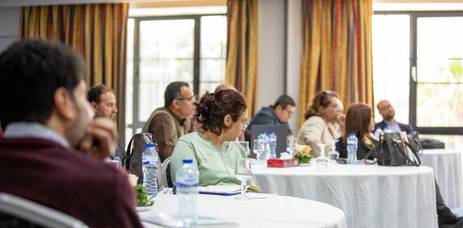 Konferenzraum mit mehreren runden Tischen. Es sitzen diverse Personen an den Tischen. Wasserflaschen und Gläser stehen bereit.
