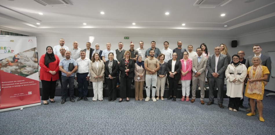 Eine große Gruppe von Menschen steht in einem Konferenzraum. Links ist ein Roll-up mit dem RECAP-Logo sichtbar.